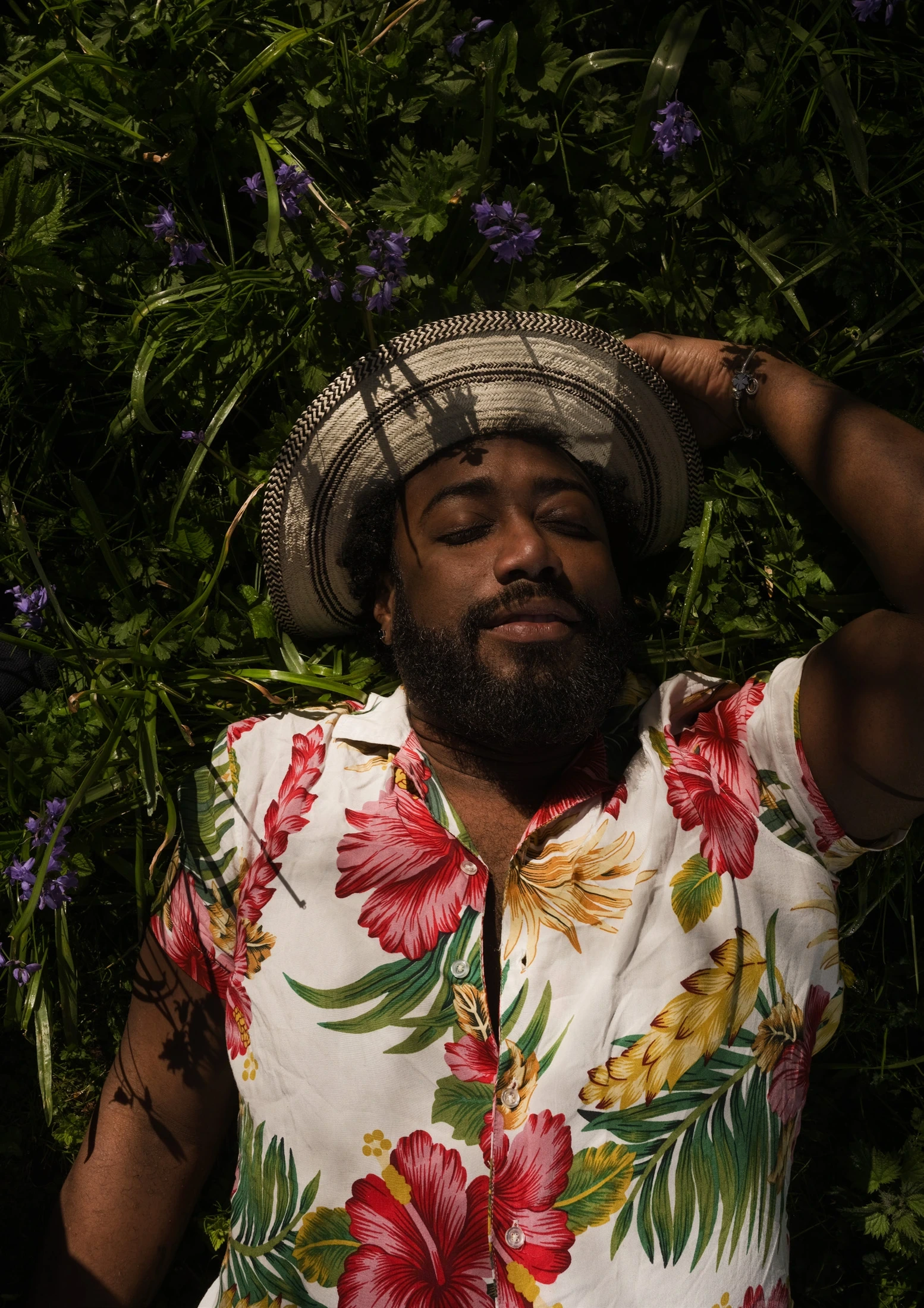 A man in a floral shirt reclining among green leaves and purple flowers