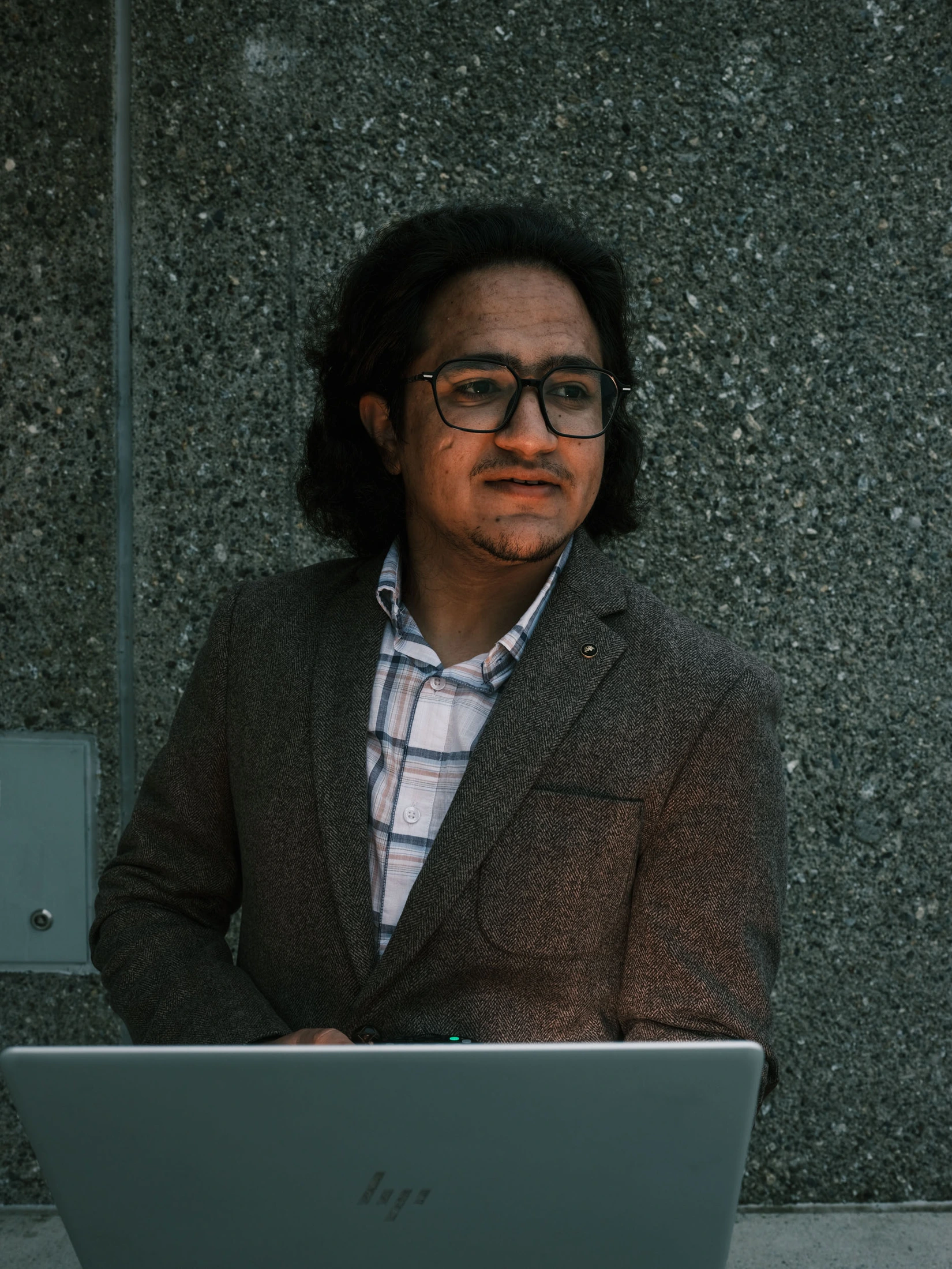 Portrait of Saeedullah wearing glasses and a brown jacket, with a laptop